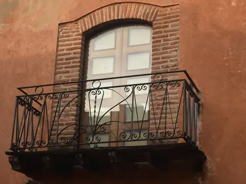 Petit balcon à garde-corps de fer forgé noir ouvragé, sous une fenêtre cintrée encadrée de briques, sur une façade ocre