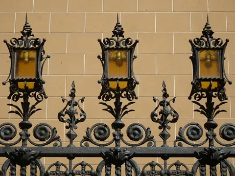 Trois lanternes en fer forgé noir à verres ambrés, posées sur une grille à fleurons et rosaces devant un mur beige