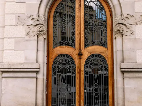 Haute porte cintrée en bois clair dont les vitrages sont protégés par des grilles de fer forgé, sous un décor de pierre sculptée