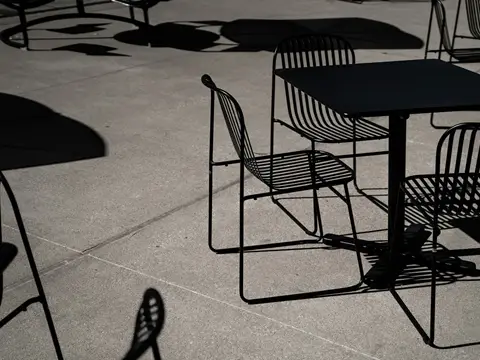 Table carrée et chaises en lames d'acier noir sur une terrasse en béton, avec de grandes ombres portées nettes en plein soleil