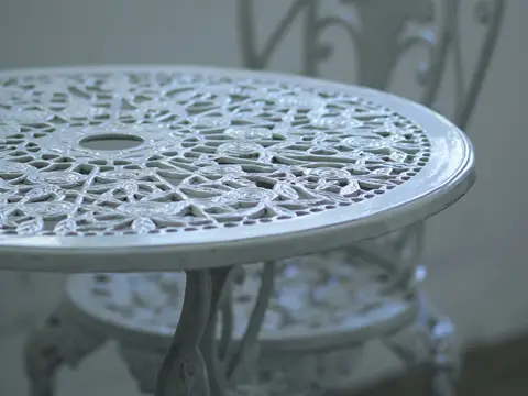 Guéridon rond en fonte blanche au plateau ajouré de roses, devant une chaise à dossier ouvragé, dans une lumière douce bleutée