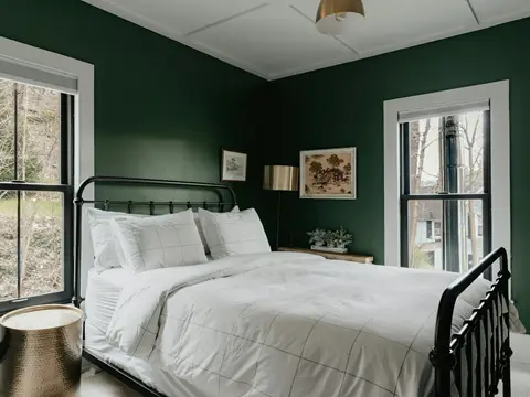 Chambre aux murs vert profond : lit en fer forgé noir, linge blanc quadrillé, lampadaire en laiton et table d'appoint en laiton martelé