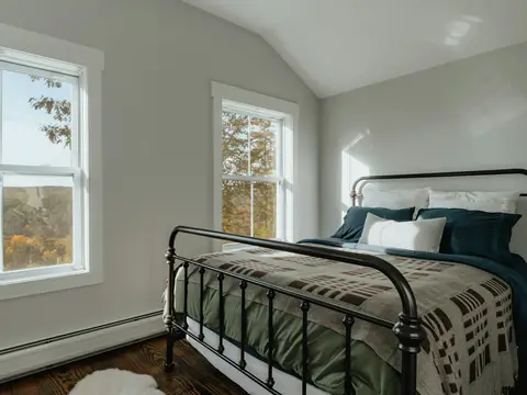 Chambre lumineuse sous combles : lit en fer noir à pommeaux, couverture à carreaux bruns, grandes fenêtres sur une forêt d'automne