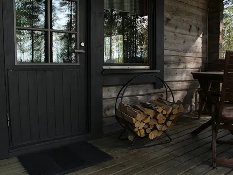 Porte-bûches en anneau d'acier noir rempli de bûches de bouleau, posé sur la terrasse en bois d'un chalet à porte noire