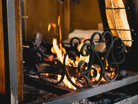 Grille ornée de volutes en fer forgé noir devant des flammes vives, dans un foyer ouvert à cadre d'acier et de bois