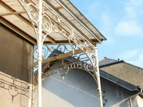 Marquise en fer forgé blanc à volutes et verre au-dessus d'une façade, sous un ciel bleu clair
