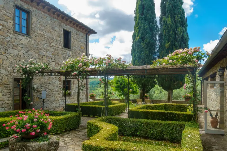 Pergola en fer forgé noir couverte de rosiers au-dessus d'un jardin de buis, devant une bastide en pierre