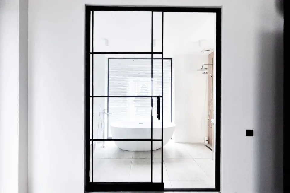 Grande porte verrière coulissante en acier noir sur un mur blanc, baignoire îlot au fond